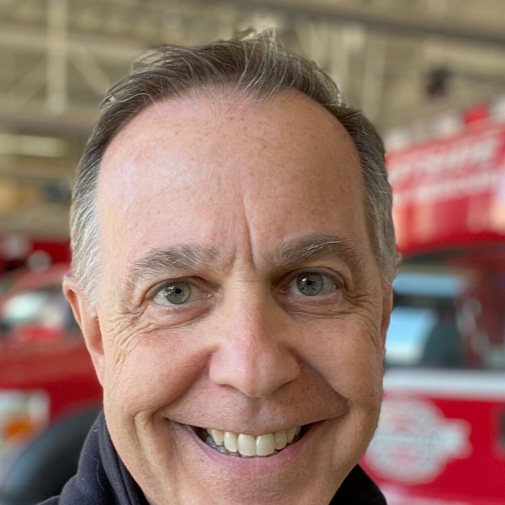 David Geller smiling in a fire station, a red fire engine behind him.